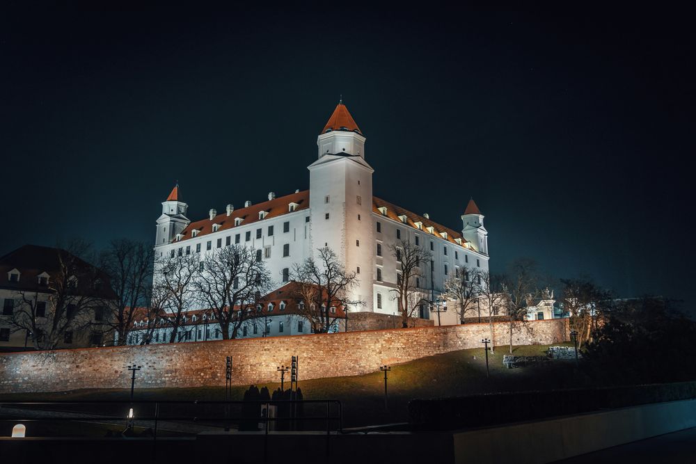 Die weiß getünchte Burg Bratislava mit ihren vier Ecktürmen und roten Dächern, angestrahlt in der Nacht über der Stadtmauer