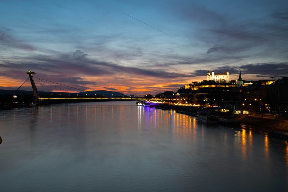 Die Donau bei Sonnenuntergang, links der UFO-Turm auf der Brücke, rechts die beleuchtete Burg, in der Mitte ein Ausflugsschiff auf dem Wasser