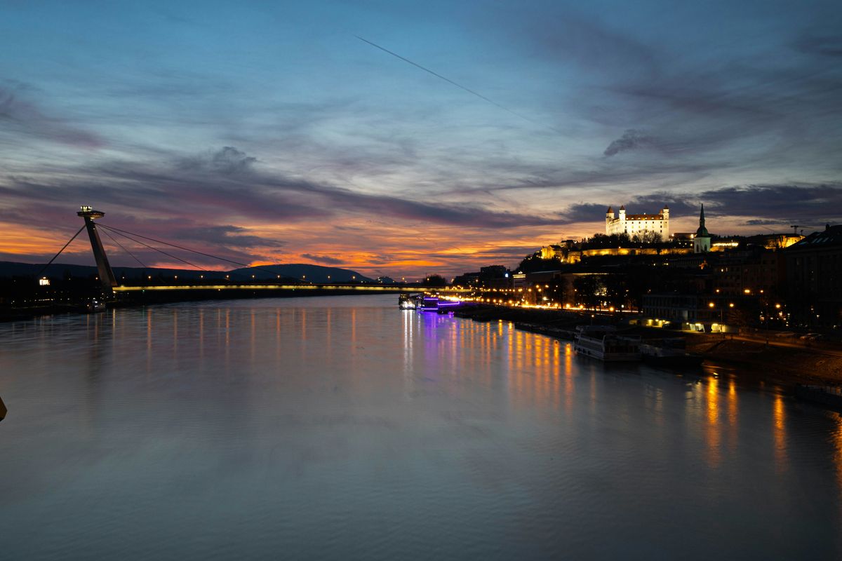 Die Donau bei Sonnenuntergang, links der UFO-Turm auf der Brücke, rechts die beleuchtete Burg, in der Mitte ein Ausflugsschiff auf dem Wasser
