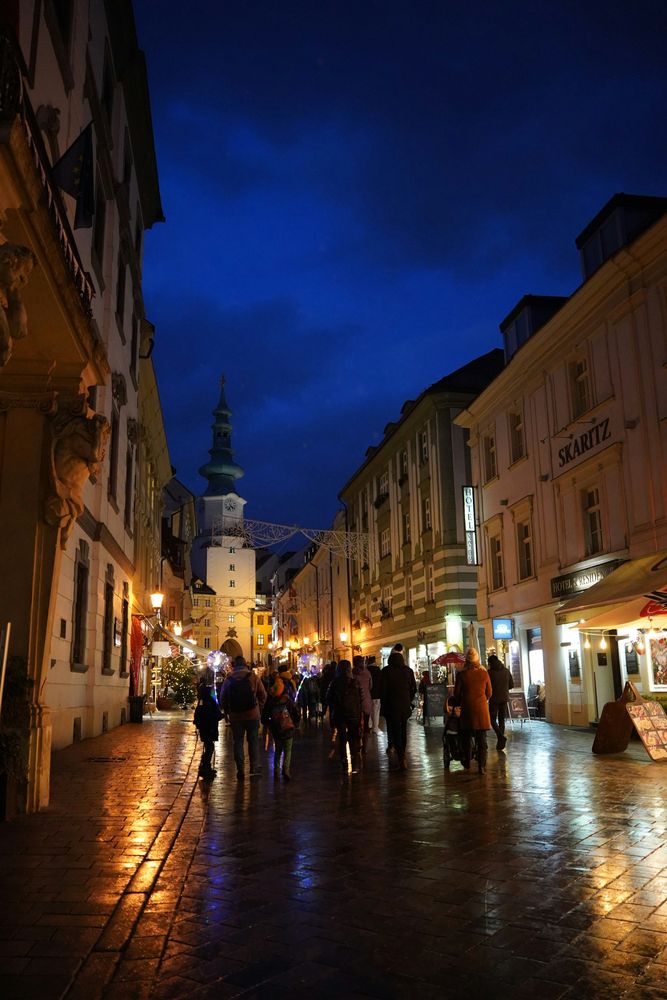 Abendliche Gasse in der Bratislavaer Altstadt mit Lichterketten über der Straße, Passanten auf nassem Kopfsteinpflaster und dem beleuchteten Michaeler Tor im Hintergrund