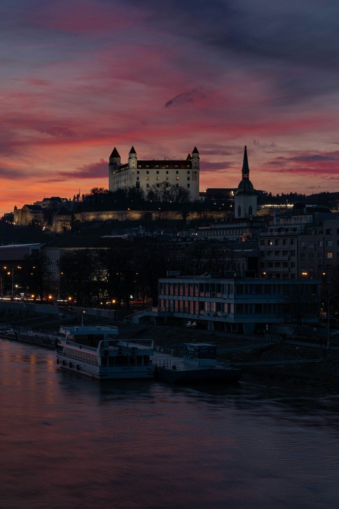 Die Burg Bratislava vor rotem Abendhimmel über der Donau, davor die Uferpromenade mit Schiffsanlegern
